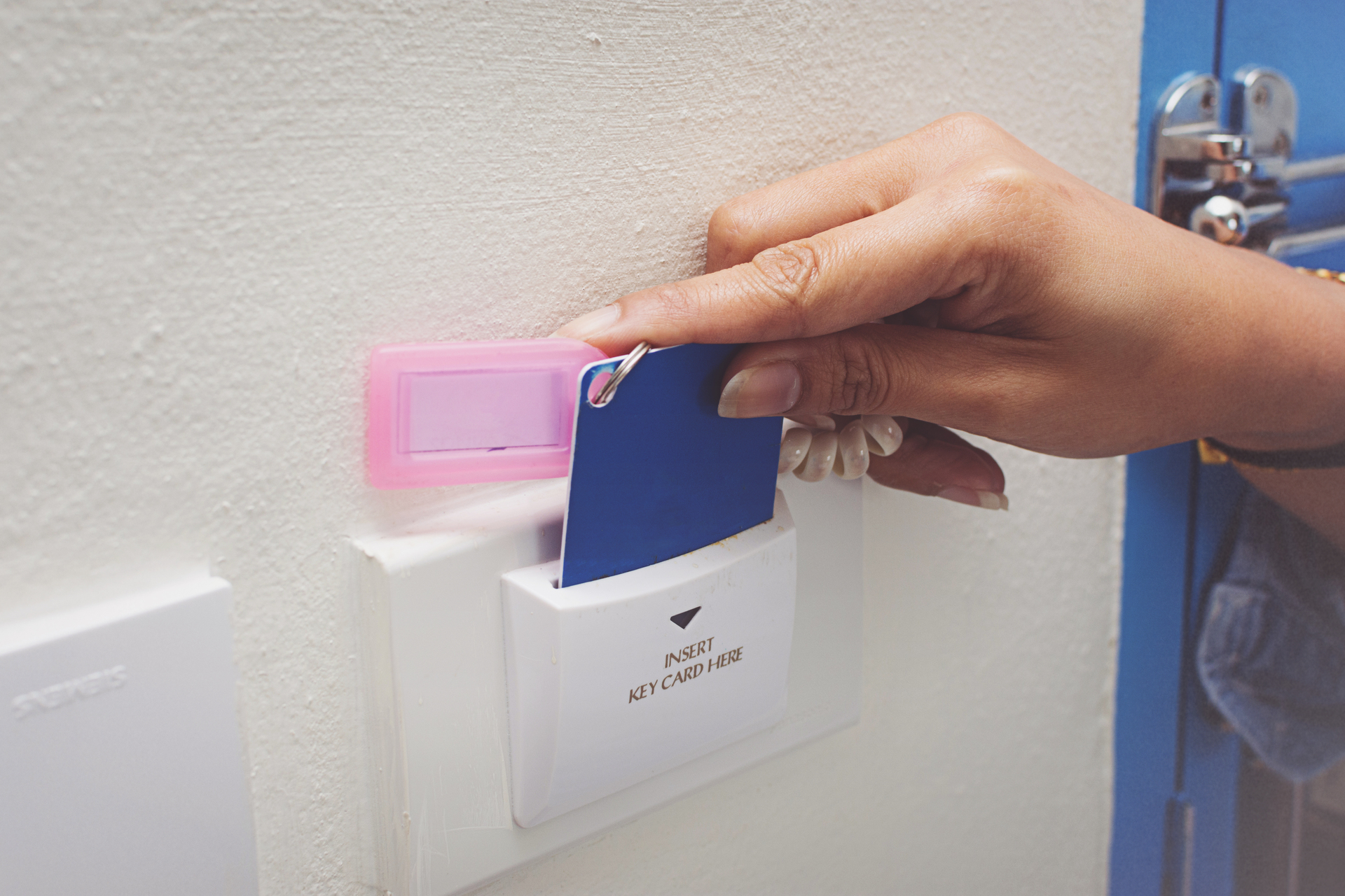 En hand håller ett blått nyckelkort framför en vit elektronisk läsare med texten "INSERT KEY CARD HERE". Läsaren är monterad på en vit vägg, och ovanför den finns en rosa plastficka. Bakgrunden visar en del av en blå dörr med en silverfärgad låsmekanism